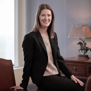 Abby Abruzzo stands with arms crossed and smiling at the camera. She has shoulder-length blonde hair, a bright smile, and a dark blazer on over a gray top.