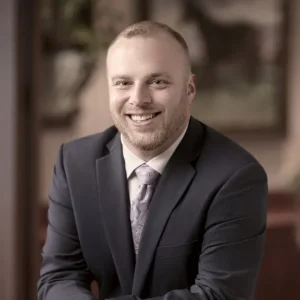 Adam Zajac is smiling at the camera. He has a buzz-cut hairstyle with a dark gray suite and gray paisley tie.