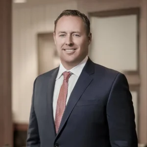 Casey Gober smiles at the camera. He is wearing a dark gray suite, red and gold spotted tie, and has short brown hair.