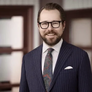 Dylan Roberts smiles at the camera. He has slicked back brown hair, black glasses, a navy pin-striped suite, and a paisley tie.