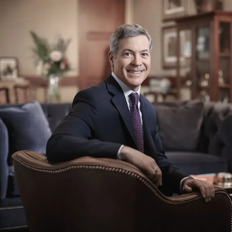 James Pruett is seated in a leather chair and is turned to face the camera. He has gray hair, with a black suite and purple speckled tie.