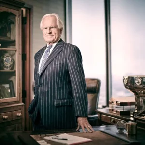 James J Drury III stands in a sharp pinstriped suit, with white hair and a stoic look on his face. His hand is laying on a desk in a classy office.