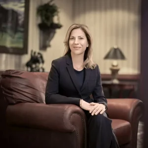 Nancy Konieczki is sitting in a large brown leather chair with her hands clasped on her crossed knees. She is wearing a black suite over a black top, with shoulder-length blonde hair.
