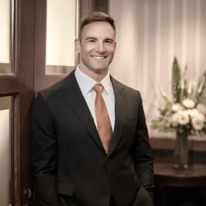 Sam Albright is wearing a black suite with a red and gold striped tie. He is smiling at the camera while leaning against a door.