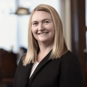 Sarah Shafarik smiles at the camera. She has shoulder-length blonde hair and is wearing a black blazer over a white top.