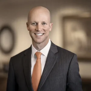 Steven Lewis smiles at the camera. He is bald, with a dark gray striped suite and red tie.