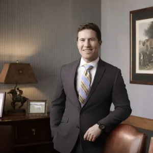 William Stanbrook stands in an office, with one arm leaning on a leather chair. He is smiling at the camera, wearing a dark gray suite with a blue and yellow striped tie.