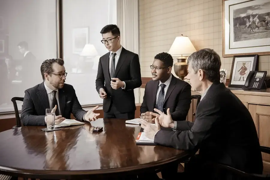 Four men in business attire hold a meeting around a round wooden table in a well-lit office with papers, glasses, and a lamp on the desk.