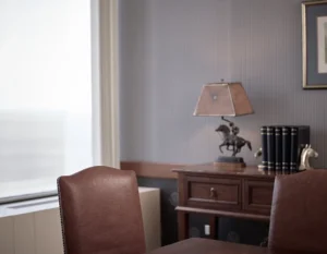 A desk with a brown lamp, a horse statue, black books, and a horse head bookend sits against a gray wall; two brown chairs are in the foreground.