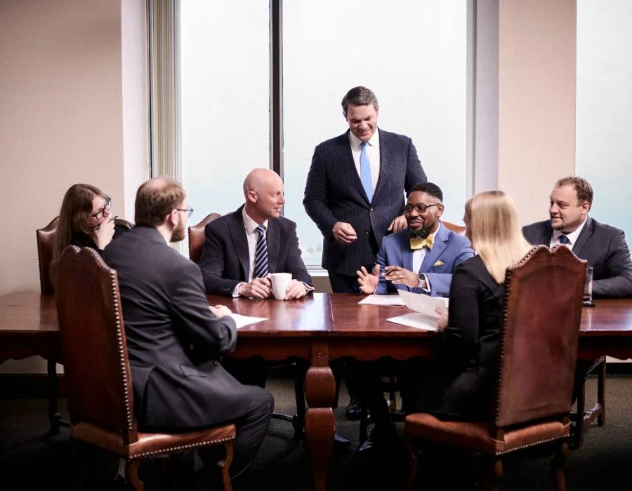 Six business professionals sit and stand around a conference table, engaged in discussion with papers in hand, in a well-lit office setting.
