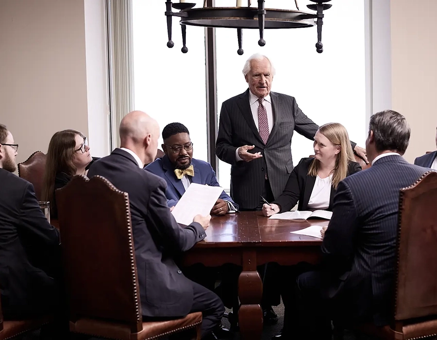 A group of business professionals sit around a conference table while an older man stands and speaks to them during a meeting.
