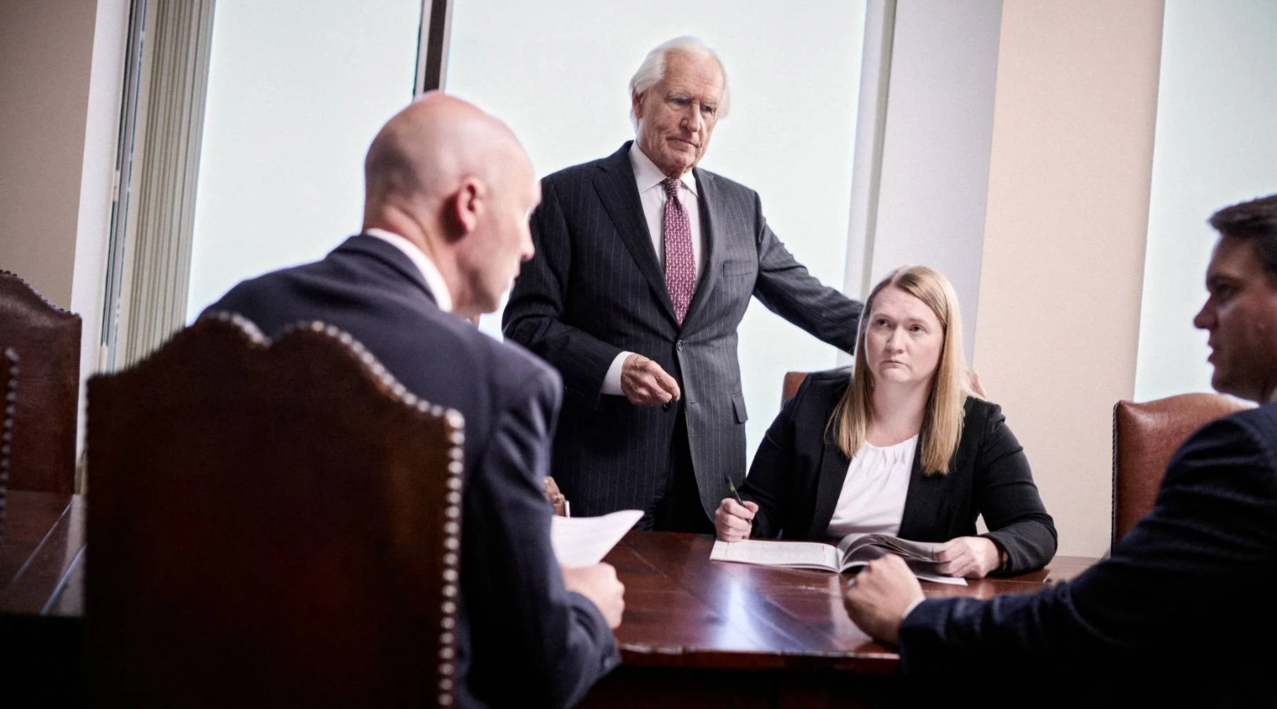 Four people in business attire sit around a conference table, with one man standing and the others seated, engaged in a meeting or discussion in a modern office setting.