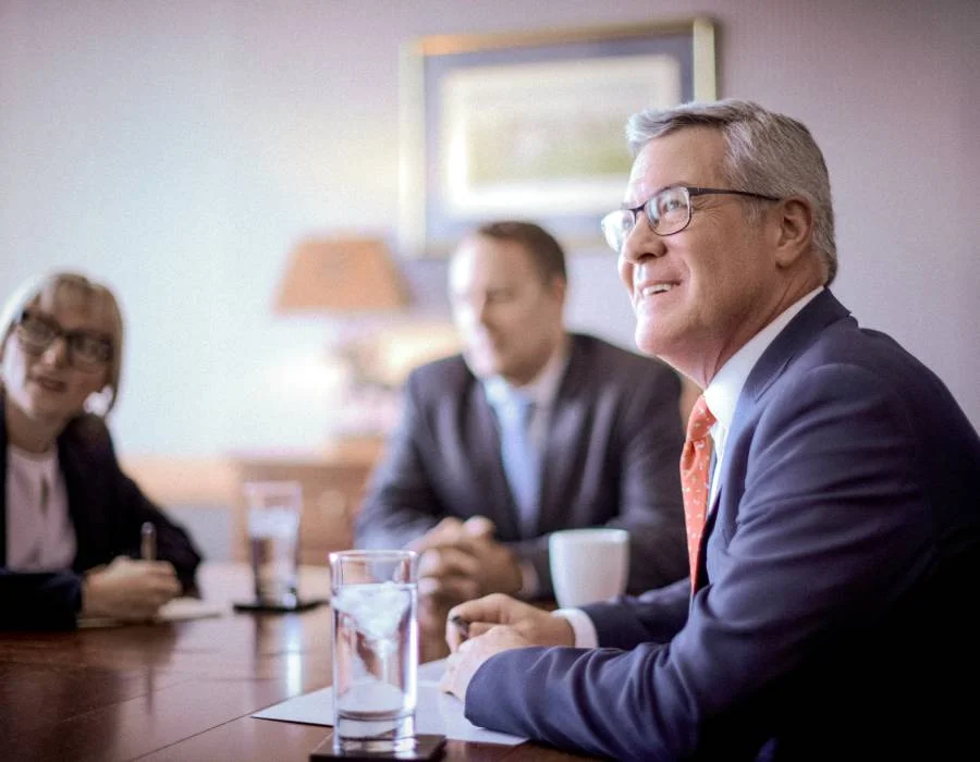 A man in a suit and glasses smiles during a meeting at a conference table, with two other blurred people seated in the background.