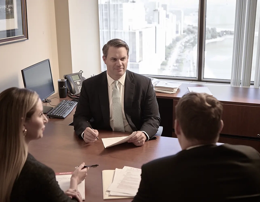 Three people in business attire sit around a desk in an office with large windows, holding documents and engaging in a meeting.