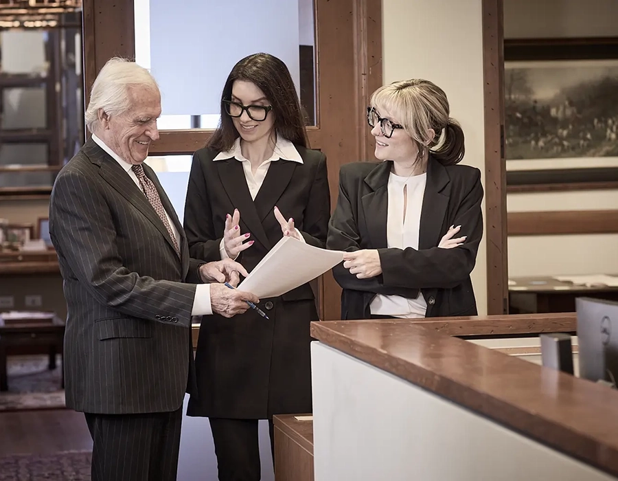 Three people in business attire stand together in an office, reviewing documents and engaged in conversation near a reception desk.