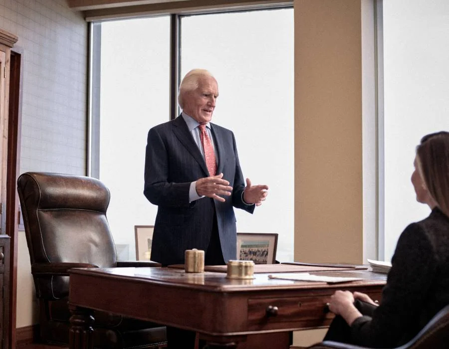 An older man in a suit speaks while standing behind a desk in a well-lit office with large windows.