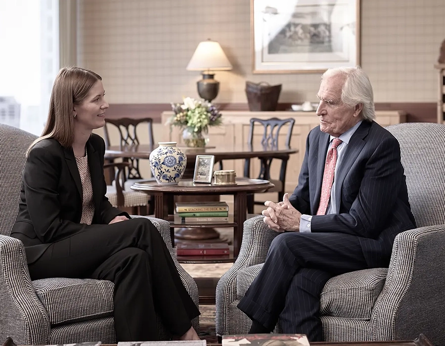 A woman and an older man in business attire sit facing each other in an office, engaged in conversation. A table with books and a vase is visible in the background.
