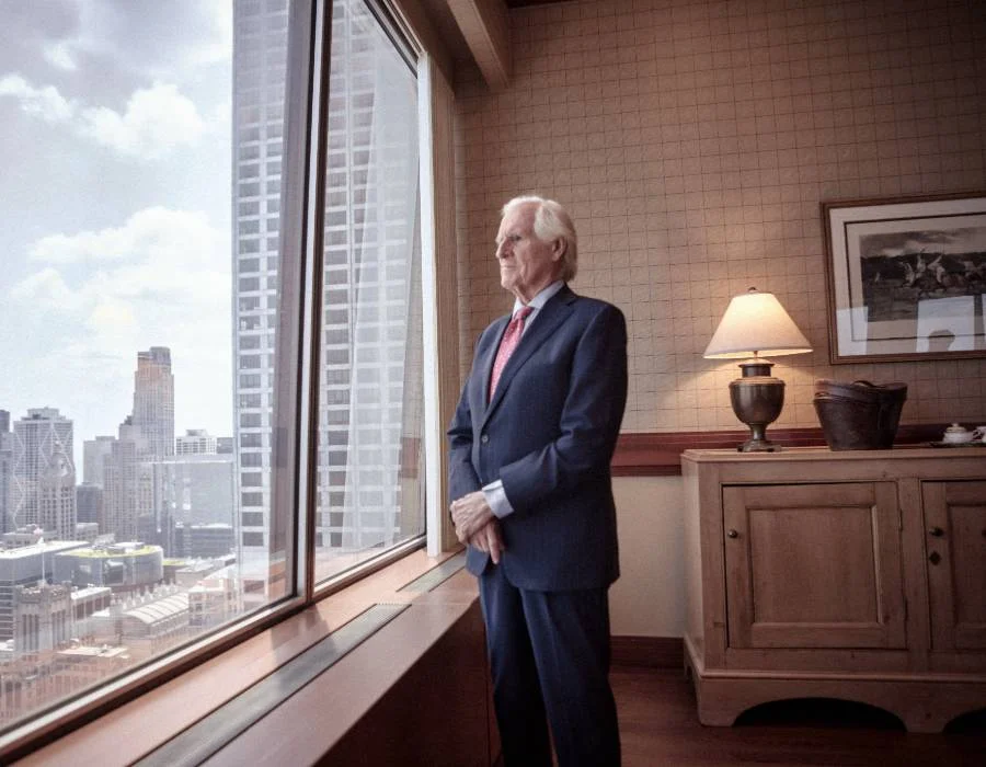 An older man in a suit stands by a window in a high-rise office, looking out over a city skyline during the day.