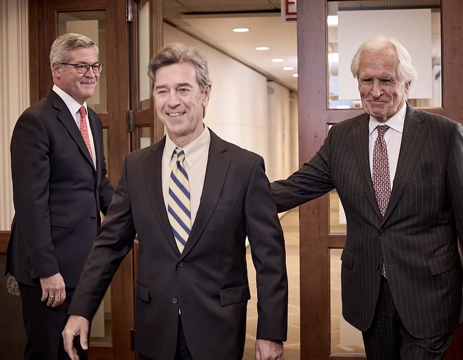 Three men in business suits stand in an office hallway; one man walks forward smiling while the other two stand near a doorway behind him.